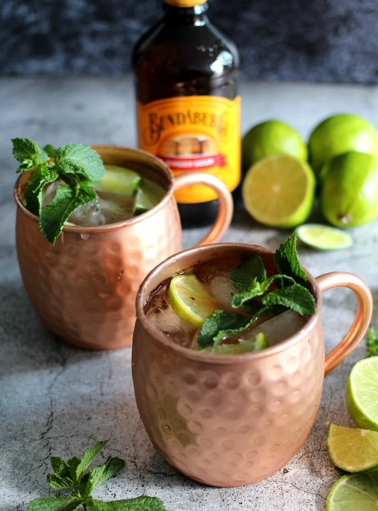 Two copper mugs filled with ice, mint leaves, and lime slices sit on a countertop. A bottle of Bundaberg ginger beer and several fresh limes, some cut, are in the background.