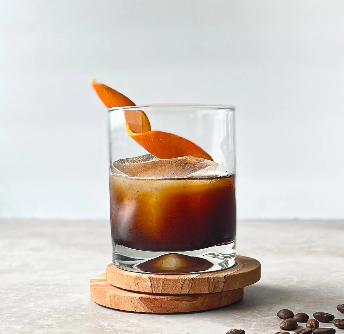 A glass of dark iced coffee with an orange peel garnish sits on round wooden coasters, with a few coffee beans scattered nearby on a light surface.