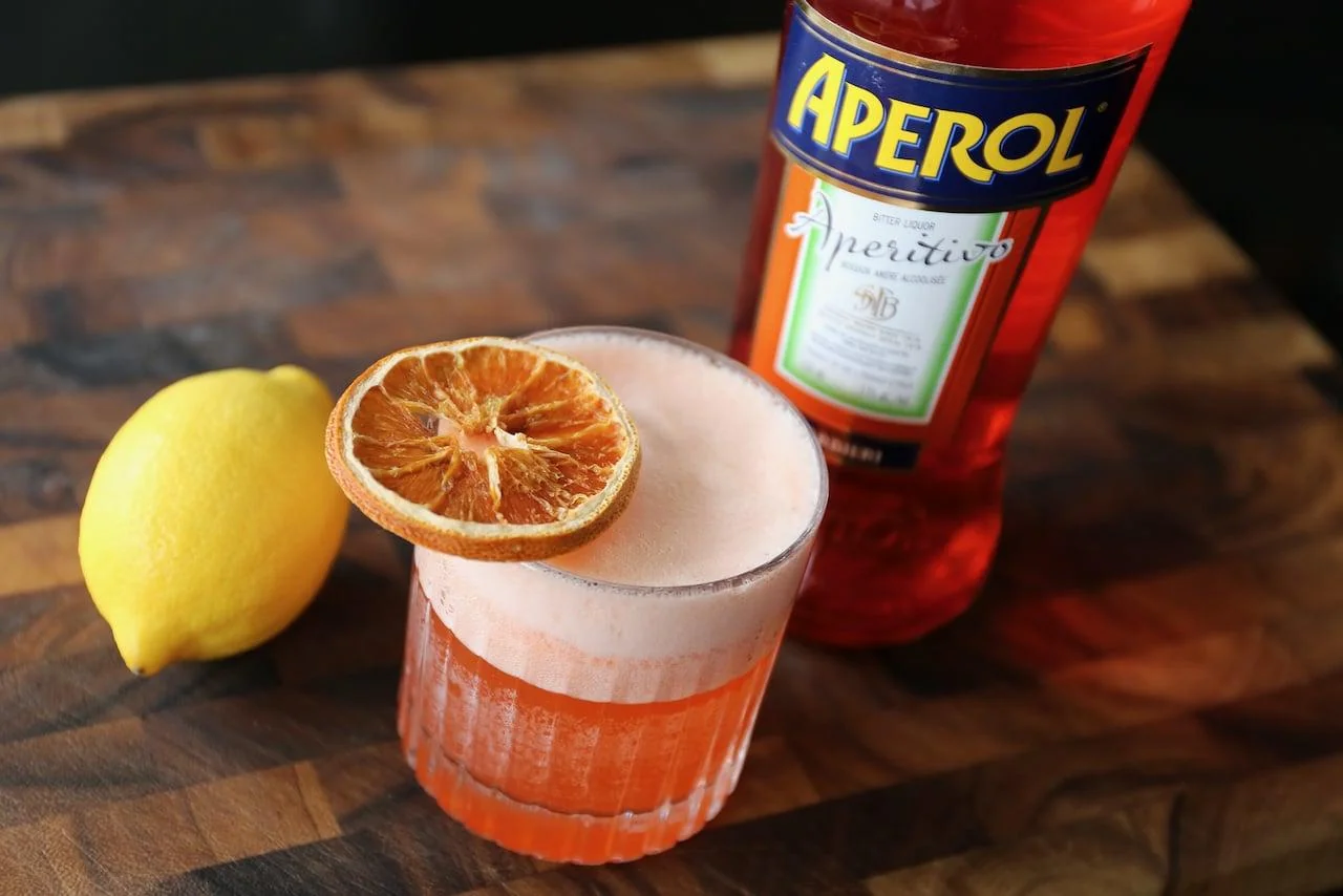 A glass of orange-colored cocktail with a frothy top and a dried orange slice garnish sits on a wooden surface next to a lemon and a bottle of Aperol.