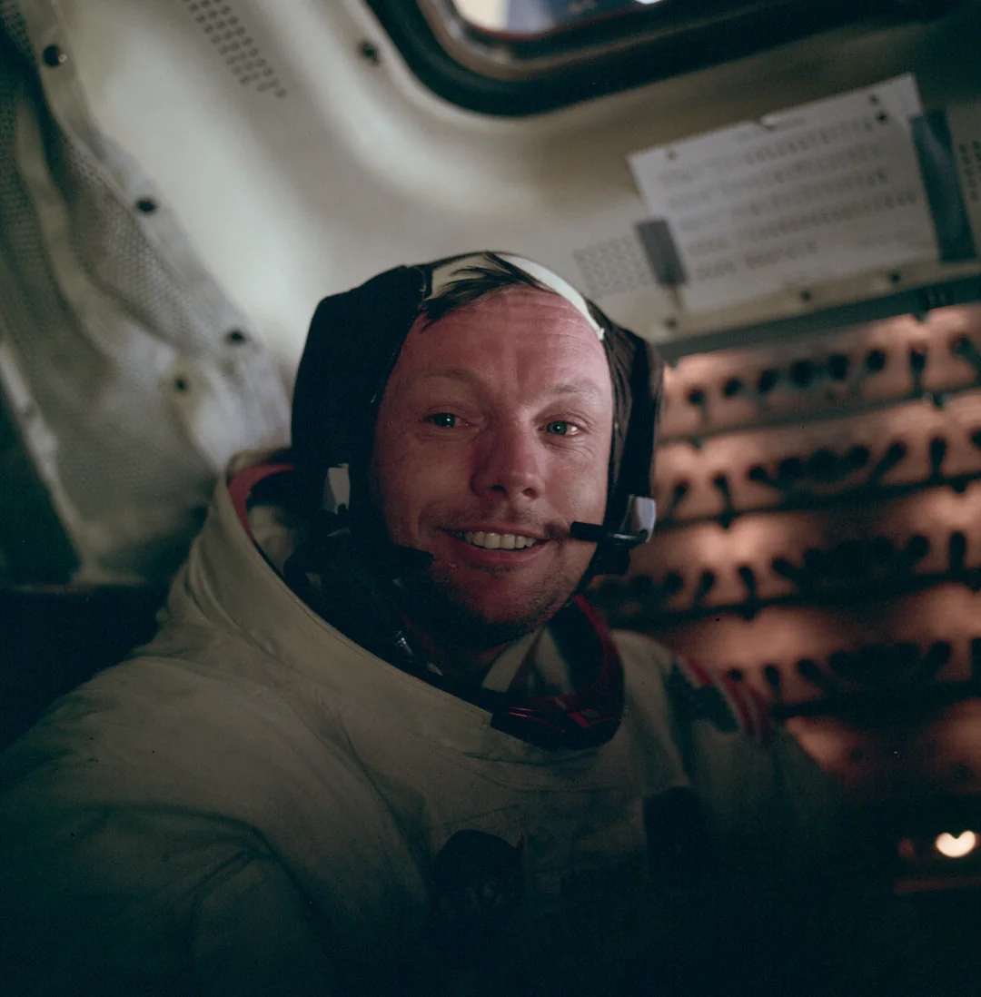 An astronaut wearing a white spacesuit and headset smiles inside a spacecraft, with control panels and a window visible in the background.