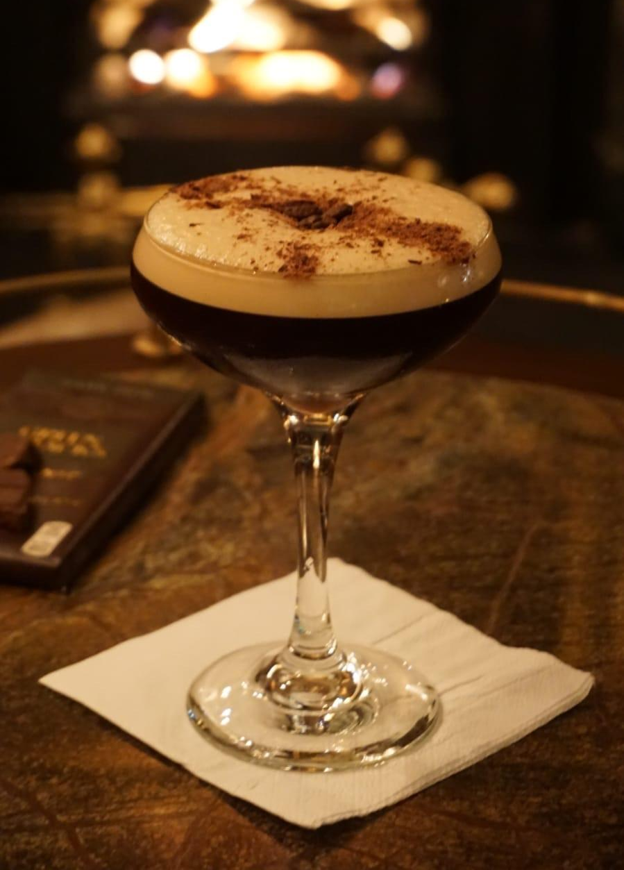 A close-up of an espresso martini in a coupe glass with a frothy top, dusted with cocoa powder, placed on a white napkin atop a wooden table, with a warm, blurred fireplace in the background.
