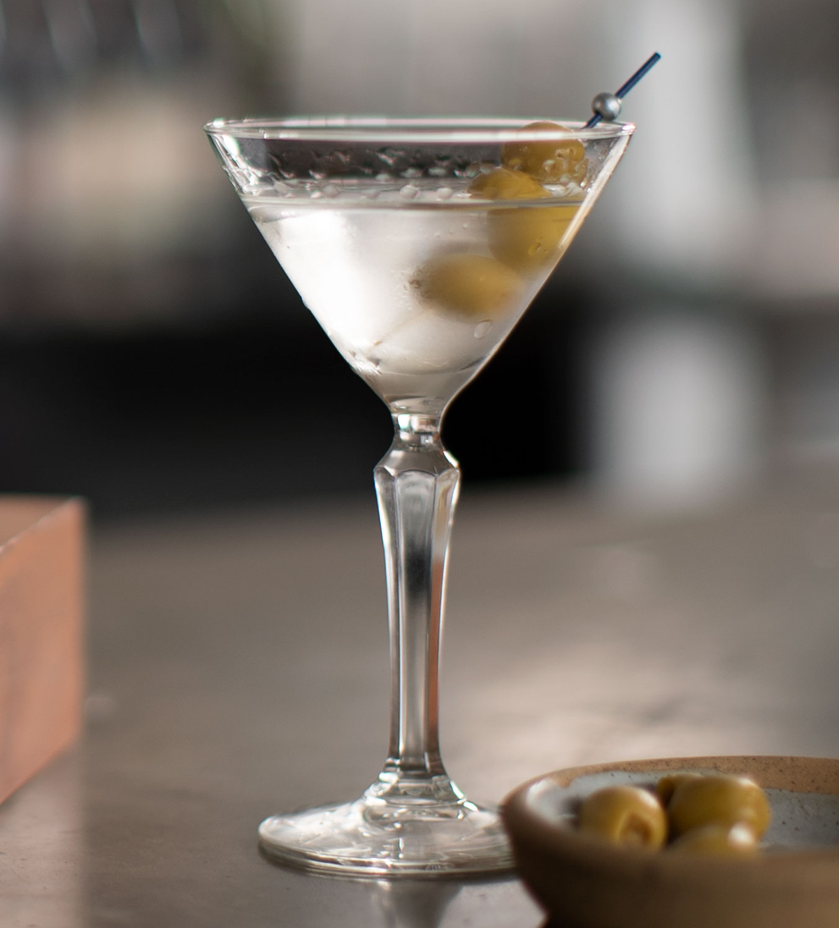 A classic martini served in a clear cocktail glass, garnished with two green olives on a pick. A small dish with more green olives is blurred in the foreground on a gray surface.