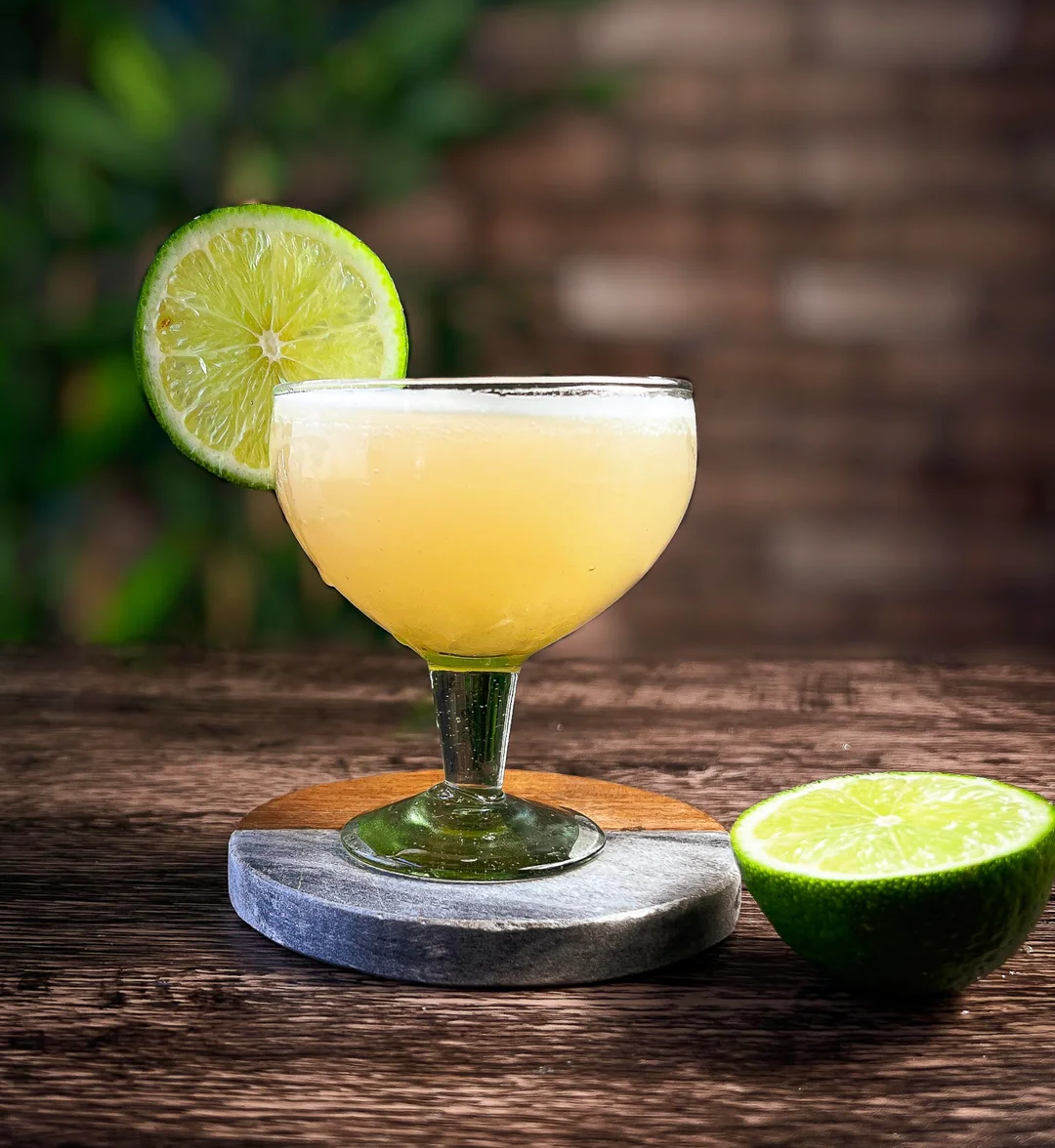 A cocktail glass filled with a yellowish drink, garnished with a slice of lime on the rim. Another half lime sits on the wooden table nearby, with a blurred green and brown background.