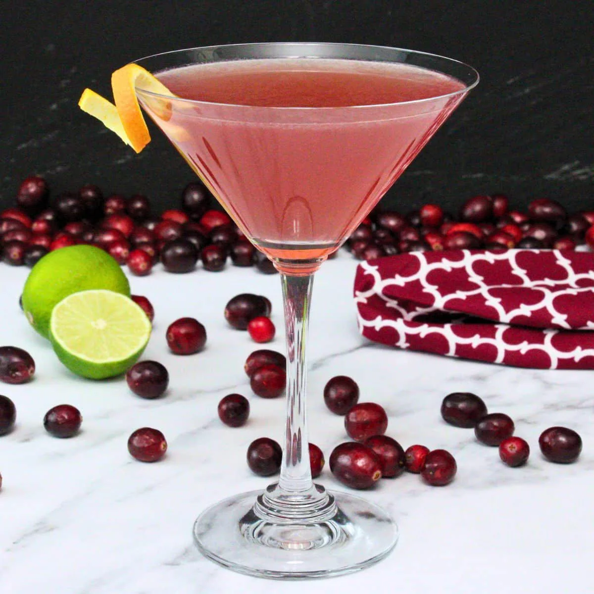 A pink cocktail in a martini glass garnished with an orange twist, surrounded by fresh cranberries and lime halves on a white surface with a red patterned cloth in the background.