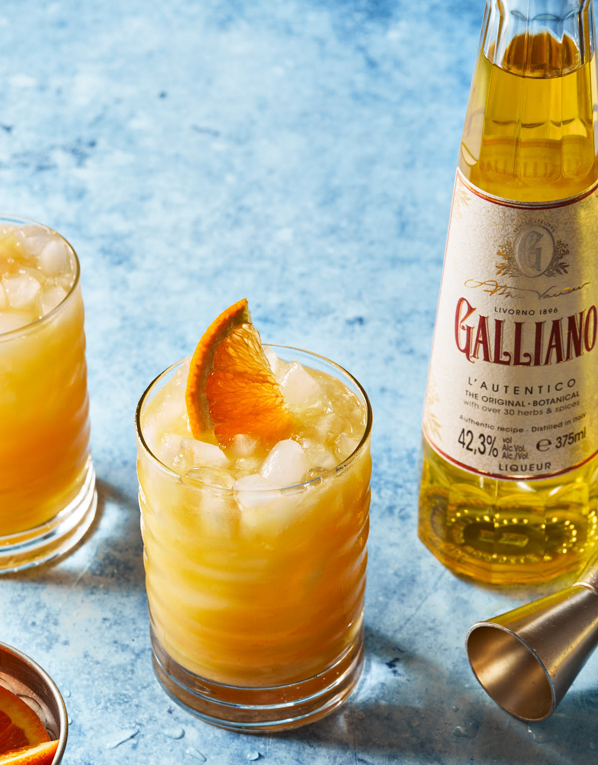 A glass of orange cocktail with ice and an orange wedge garnish sits next to a Galliano liqueur bottle on a blue surface. Another glass and citrus slices are partially visible.