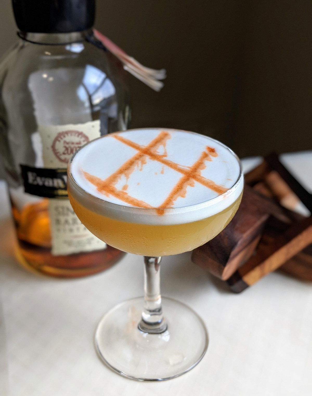 A cocktail in a coupe glass with a foamy top, garnished with orange bitters in a grid pattern, sits beside a partially visible bottle of Evan Williams bourbon and some wooden coasters.