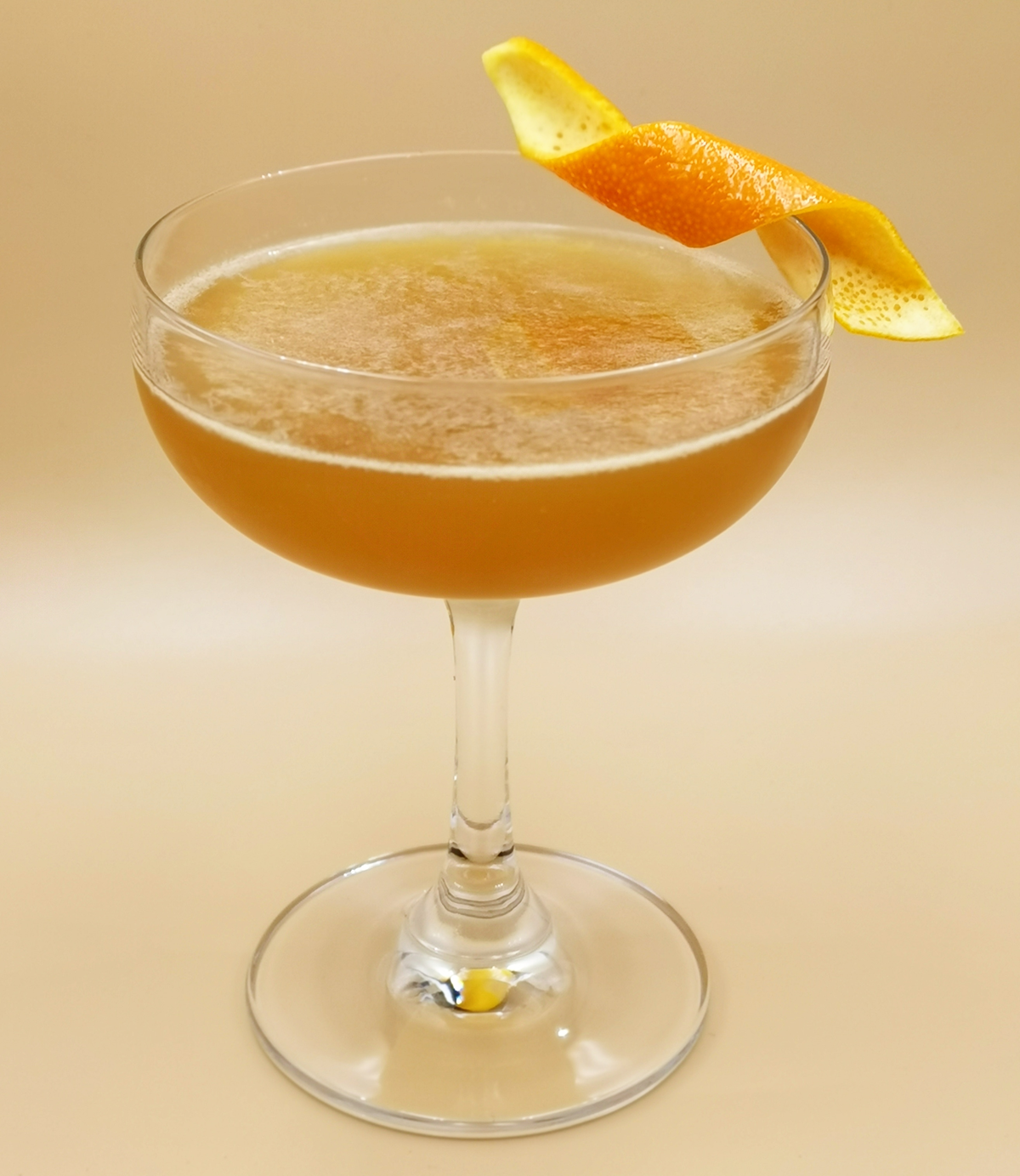 A coupe glass filled with a light brown cocktail, garnished with a twisted citrus peel on the rim, against a beige background.
