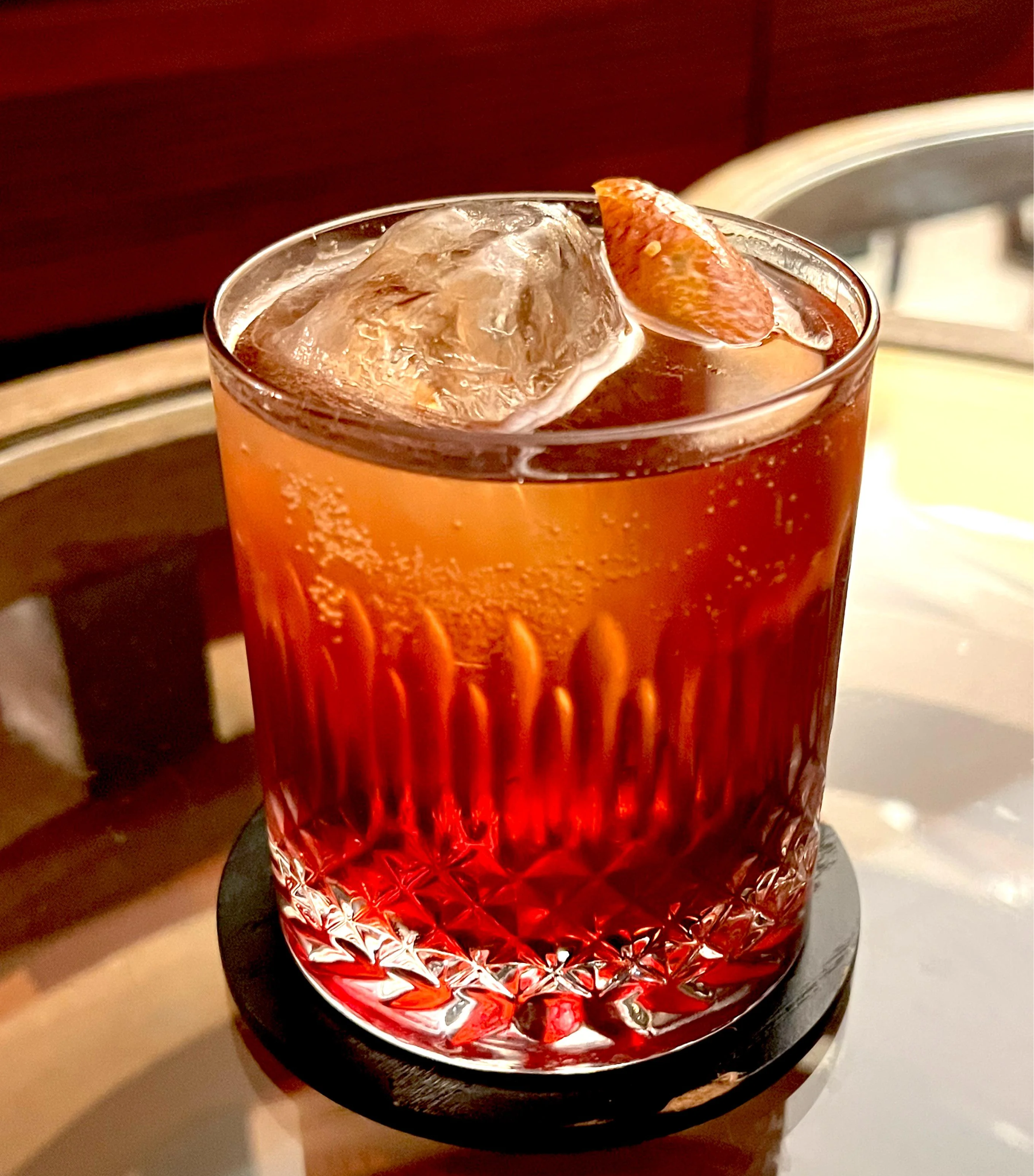 A glass tumbler filled with a red cocktail, topped with ice cubes and a slice of citrus, sits on a dark coaster on a reflective surface.