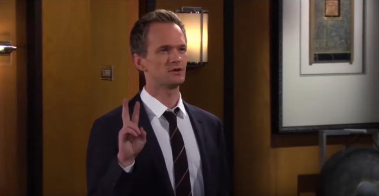 A man in a suit stands indoors, holding up two fingers as he speaks. The background shows a framed picture on the wall and part of a lamp.