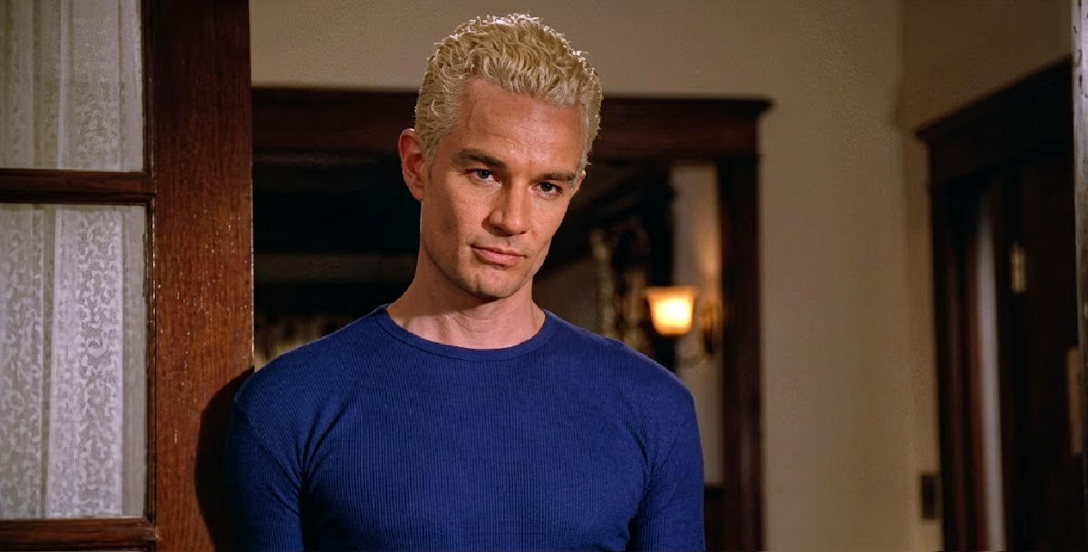 A man with short, bleached blond hair and a serious expression leans against a doorframe, wearing a fitted blue long-sleeve shirt in a warmly lit room with wooden trim.