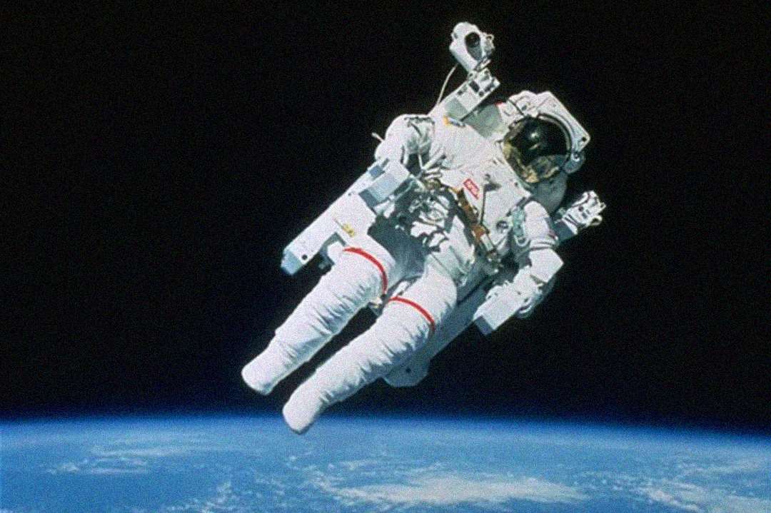 An astronaut in a white spacesuit floats above Earth, with the blue curve of the planet visible below and the blackness of space in the background.