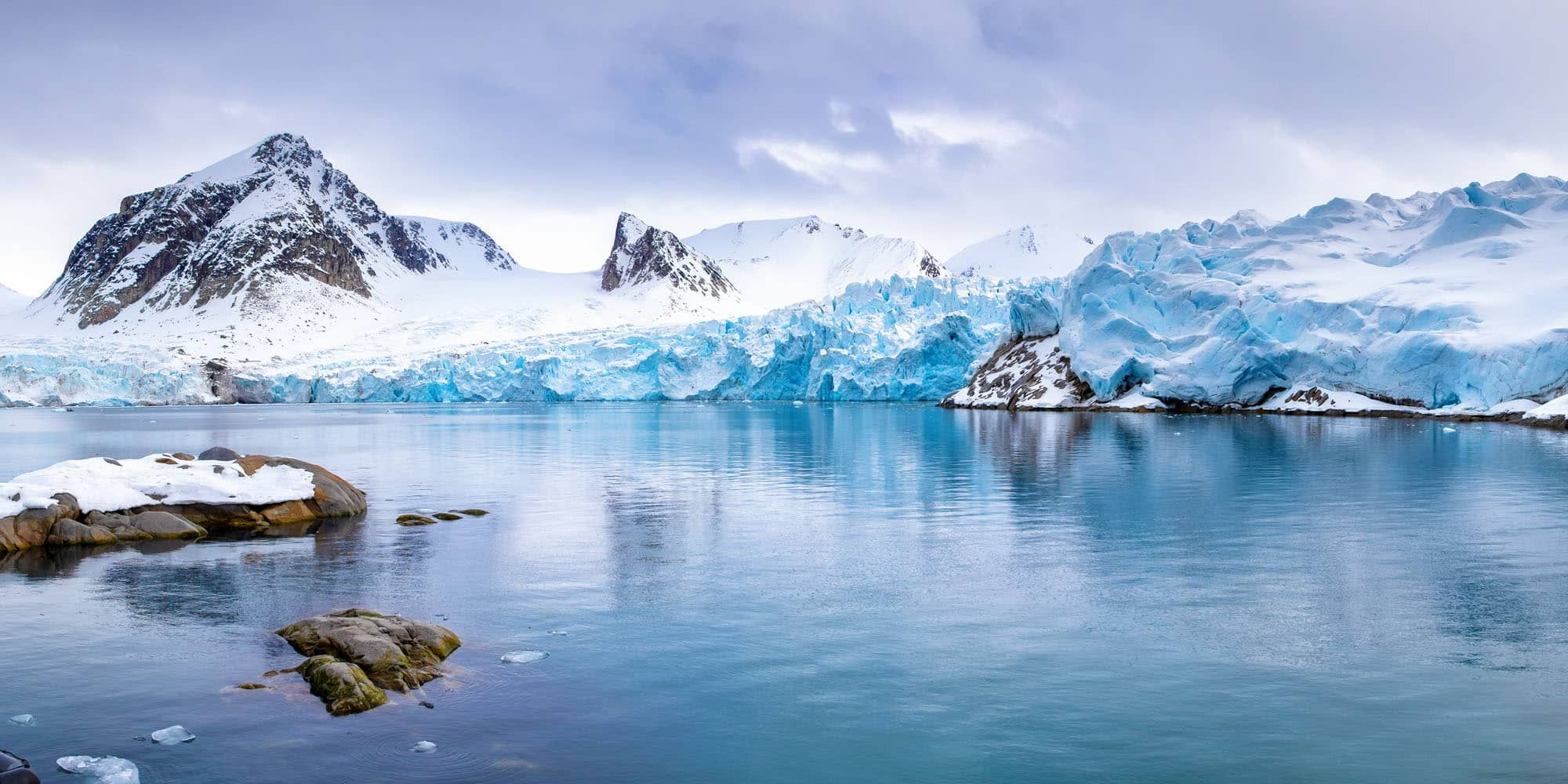 Snow-covered mountains and glaciers reflected in a calm, icy blue body of water under a cloudy sky. Rocky outcrops partially covered in snow are visible in the foreground.