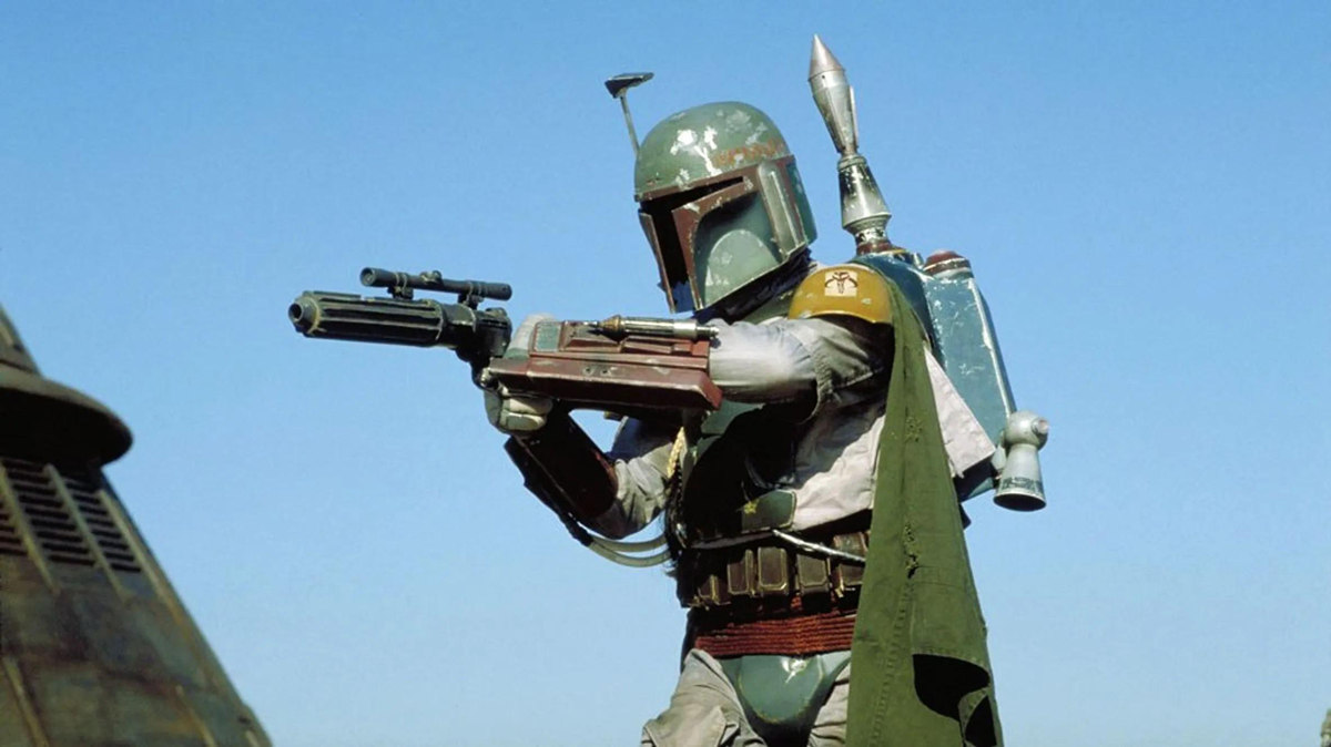 A person in green armor and a helmet with a visor, holding a blaster rifle, stands outdoors under a clear blue sky. They wear a jetpack and cape, resembling a sci-fi bounty hunter from the Star Wars universe.
