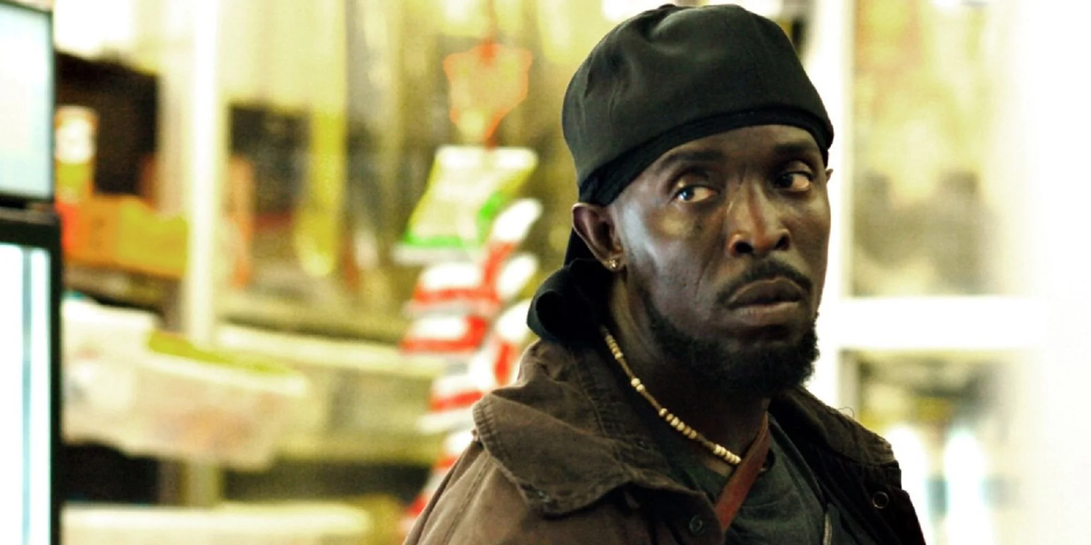 A man wearing a black backward cap and brown jacket stands indoors, looking to the side with a serious expression. Shelves with various products are blurred in the background.