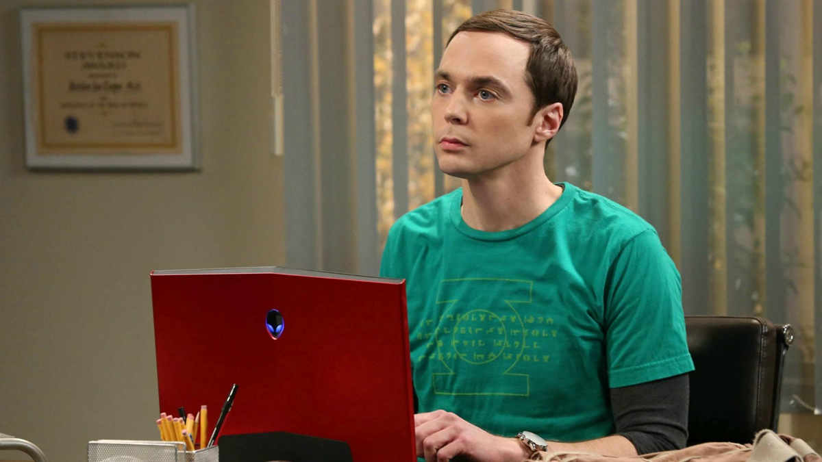 A man with short brown hair wearing a green T-shirt sits at a desk with a red laptop. He appears focused, surrounded by office supplies, with a certificate and blinds in the background.