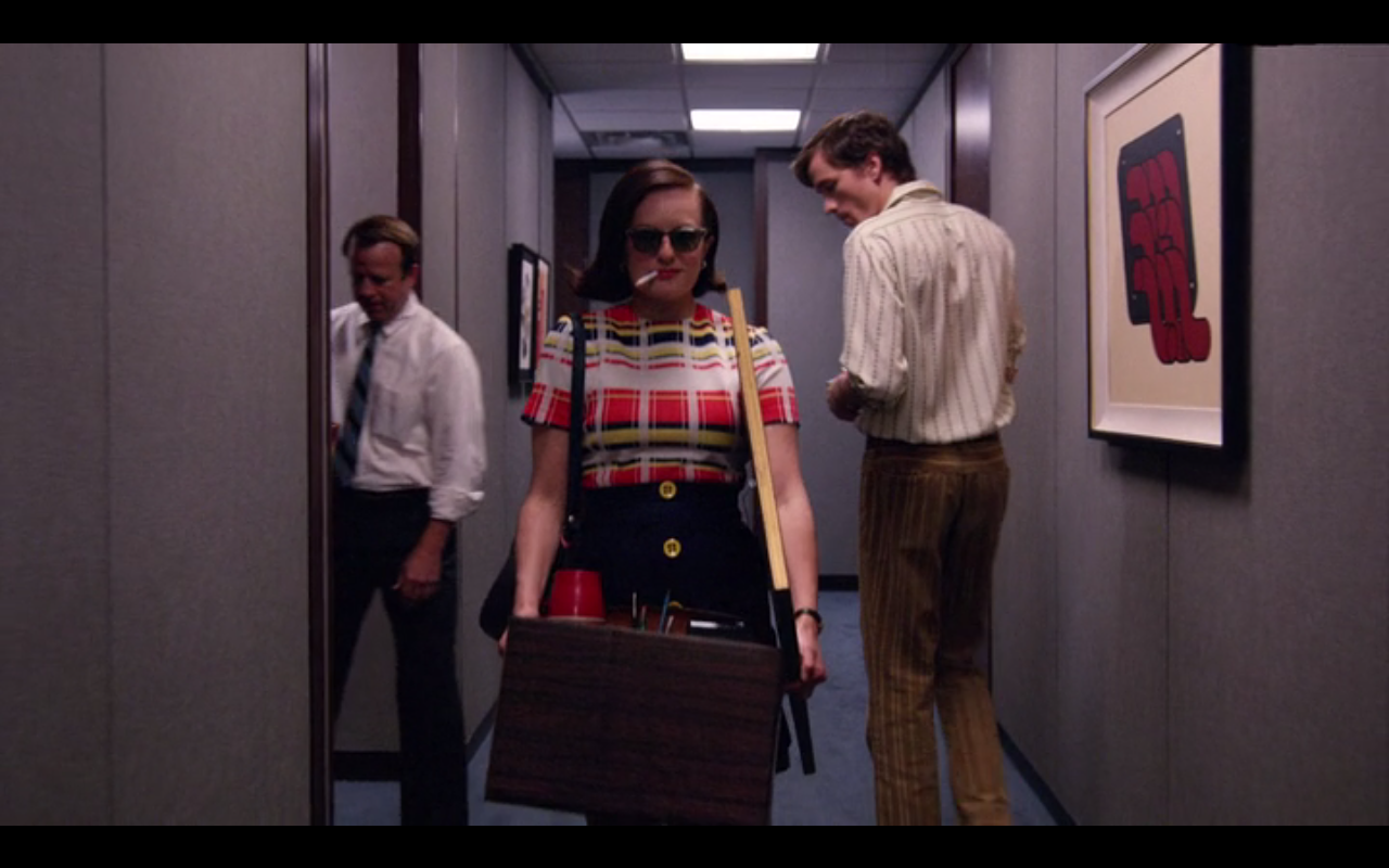 A woman in sunglasses and a plaid top confidently walks down an office hallway carrying a box of belongings, while two men in office attire stand behind her amid framed artwork on the walls.