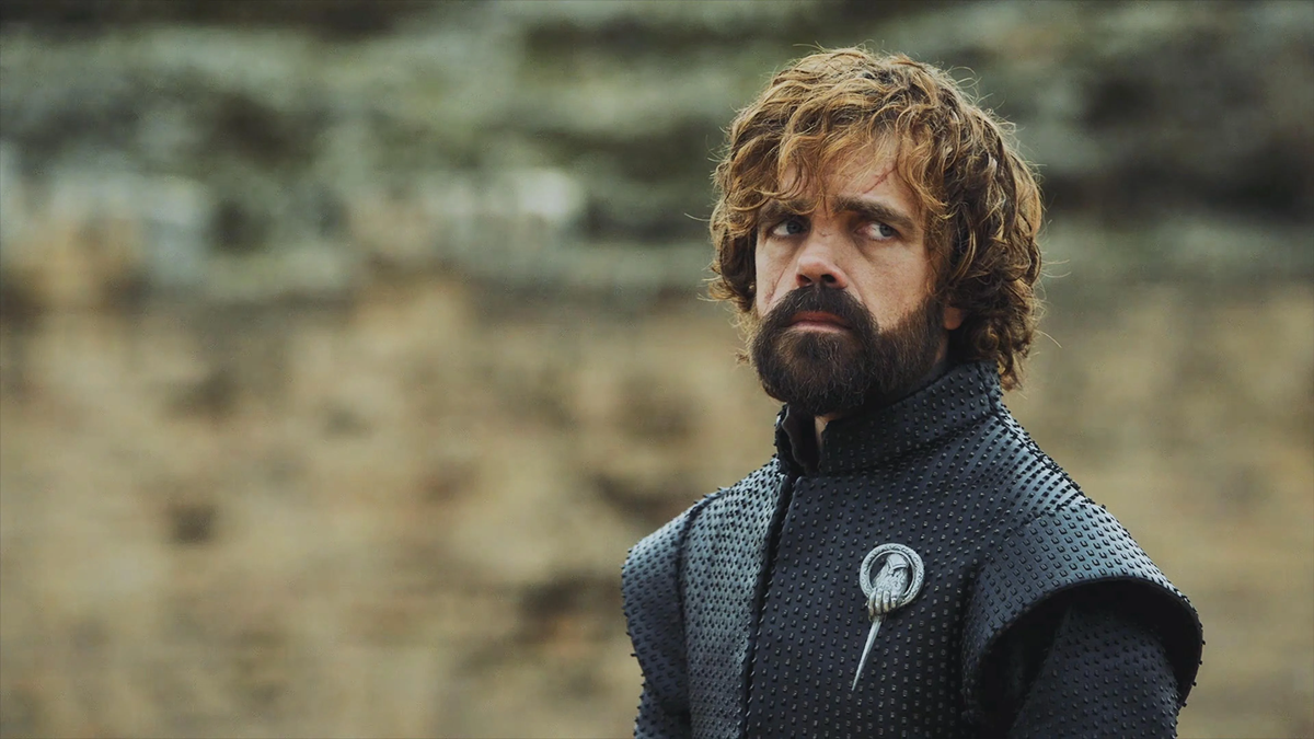 A man with curly brown hair and a beard, wearing a dark textured jacket with a silver pin, stands outdoors against a blurred stone background, looking serious.