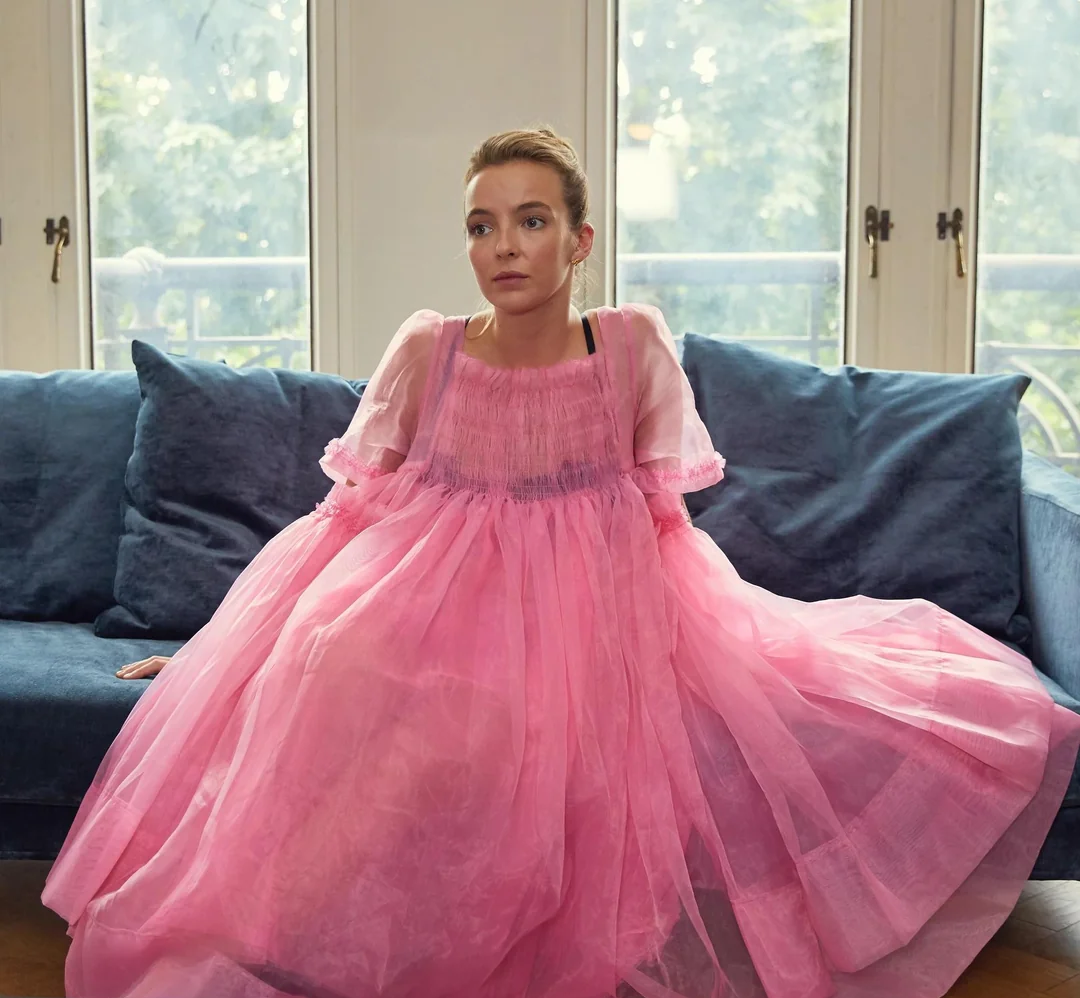 A woman in a flowing, sheer pink dress sits on a blue sofa in front of large windows with greenery outside. She looks slightly upward with a thoughtful expression.