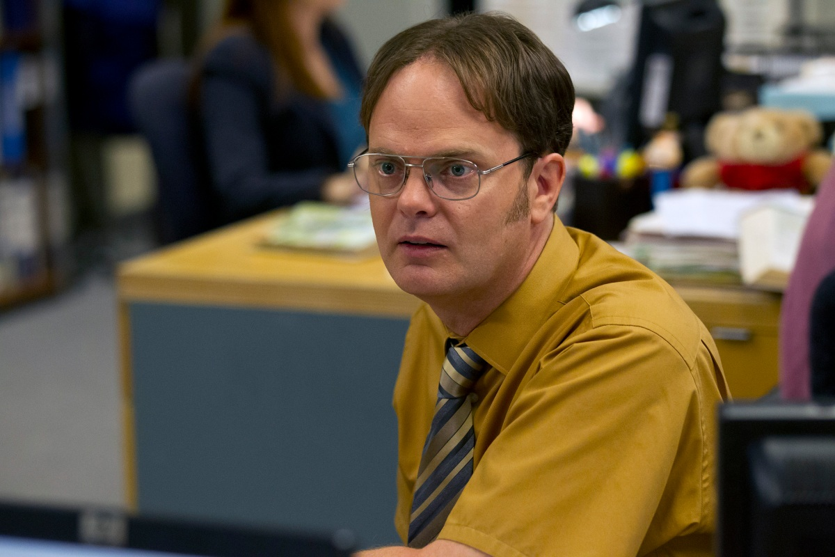 A man with glasses and short brown hair, wearing a mustard yellow shirt and striped tie, sits at an office desk looking slightly surprised. An office environment with papers and blurred figures is in the background.