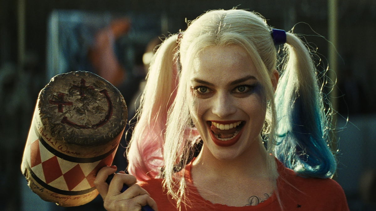 A woman with pale skin, smeared makeup, and blonde pigtails dyed pink and blue smiles widely, holding a large wooden mallet decorated with a smiley face.