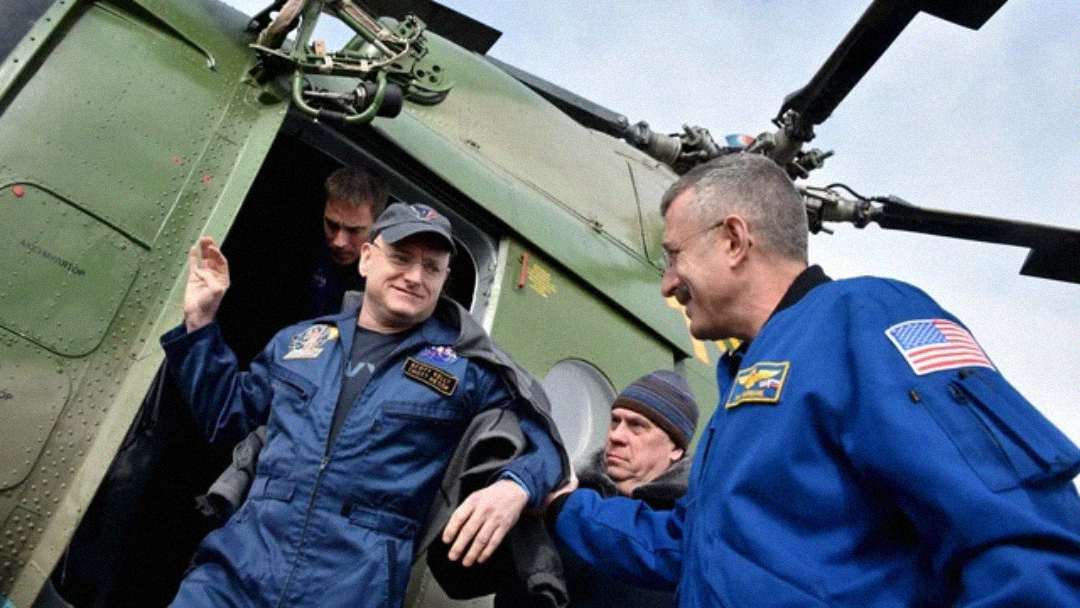 Astronauts in blue flight suits, some with NASA patches, exit a green helicopter. One astronaut is stepping out with a smile, assisted by another person as helicopter blades are visible above.