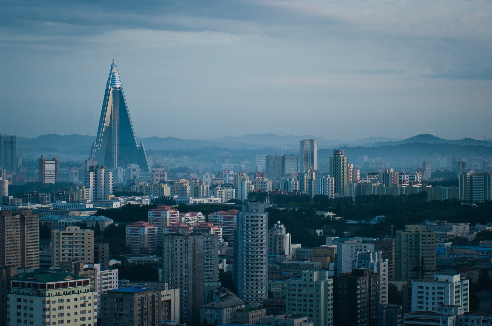 A cityscape with many high-rise buildings and a large, triangular skyscraper dominating the skyline, set against distant mountains under a cloudy sky.