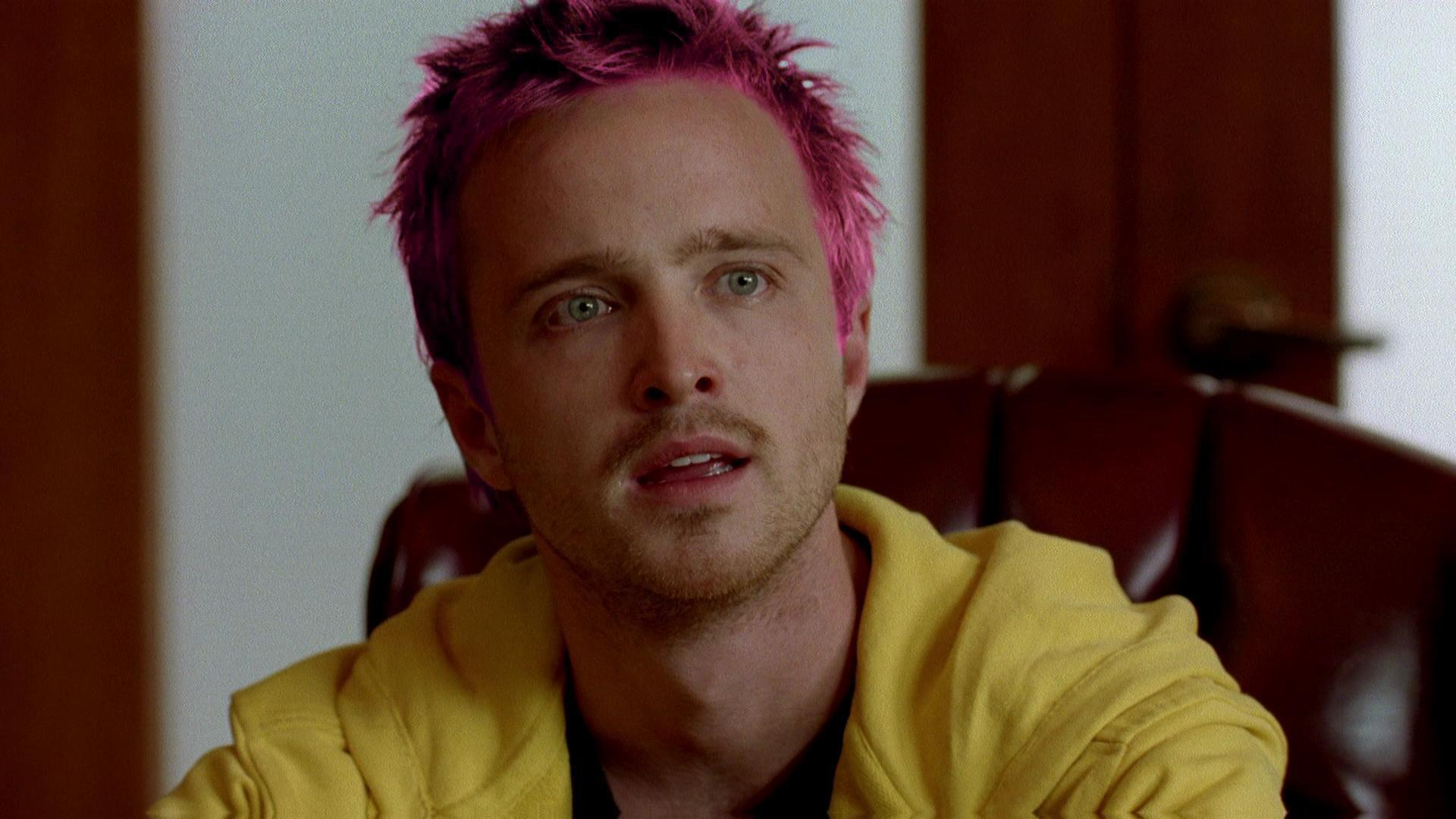 Jesse Pinkman with bright pink hair, wearing a yellow hoodie, looks off to the side with a concerned expression.