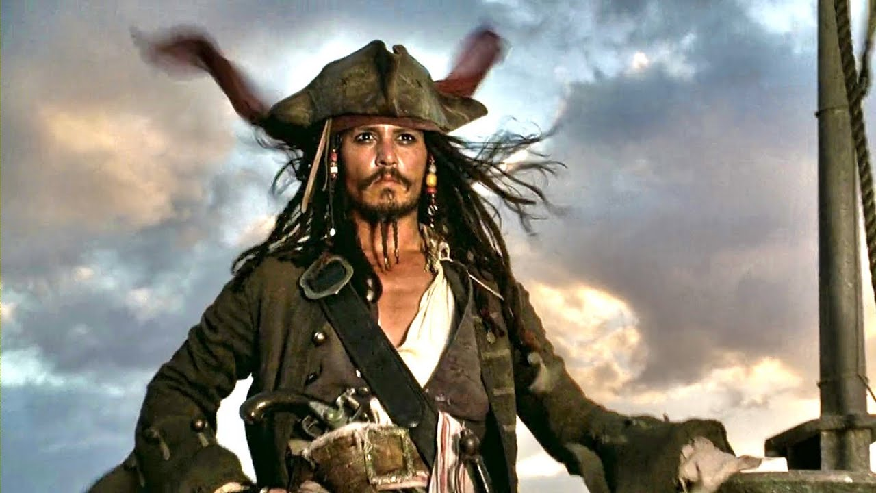 A pirate with long hair, a tricorn hat, and beaded braids stands on a ship's deck at sunset, with the sky and clouds in the background.