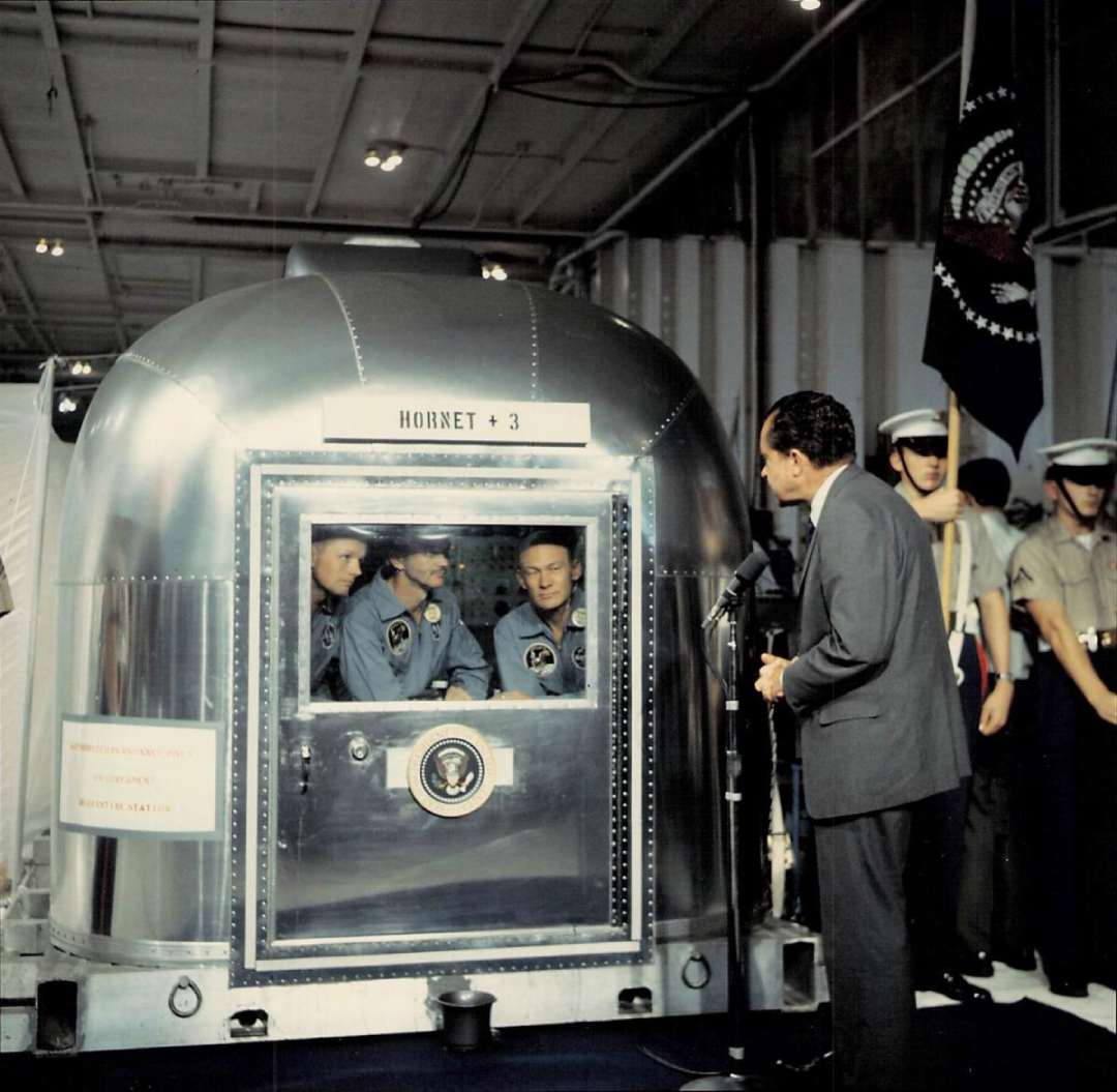 President Richard Nixon speaks to Apollo 11 astronauts, who are inside a quarantine chamber labeled “Hornet + 3,” with security and military personnel standing nearby.