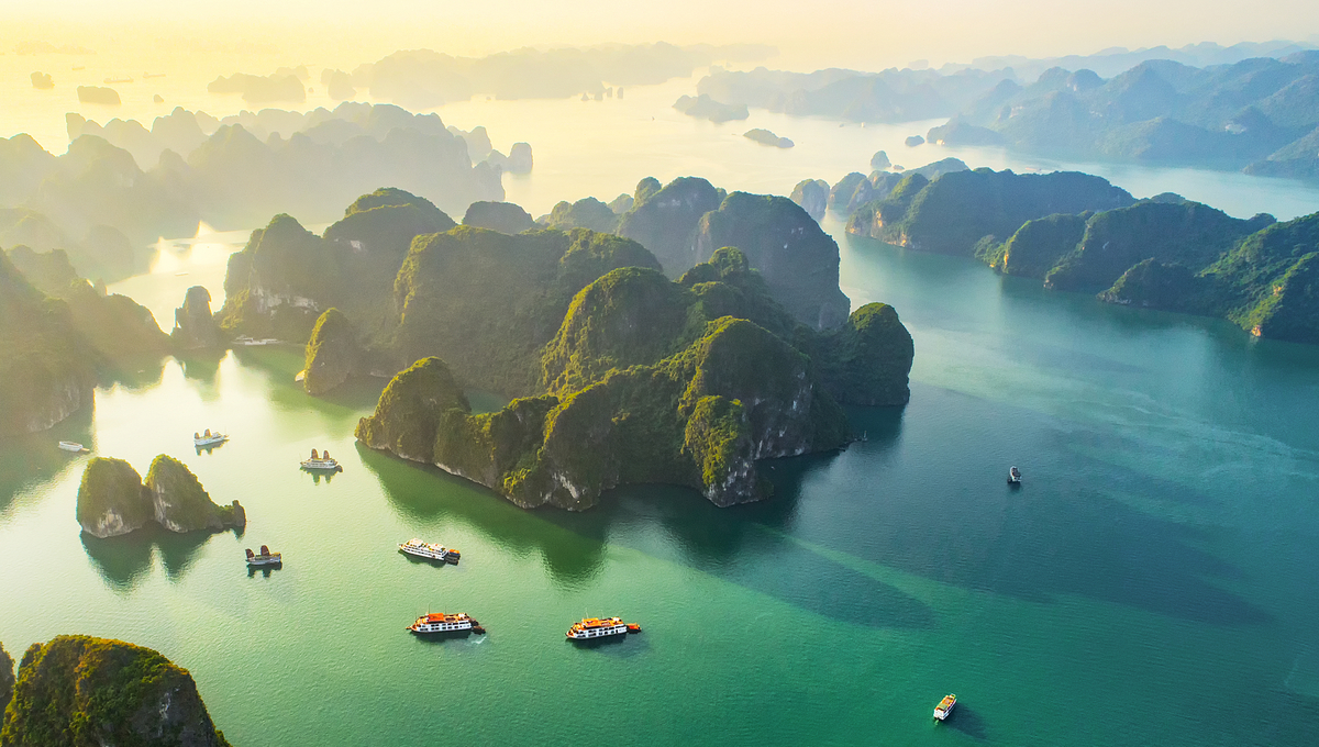 Aerial view of emerald-green water dotted with numerous lush, rocky islands and several boats cruising in the tranquil bay, with misty mountains in the background under soft sunlight.
