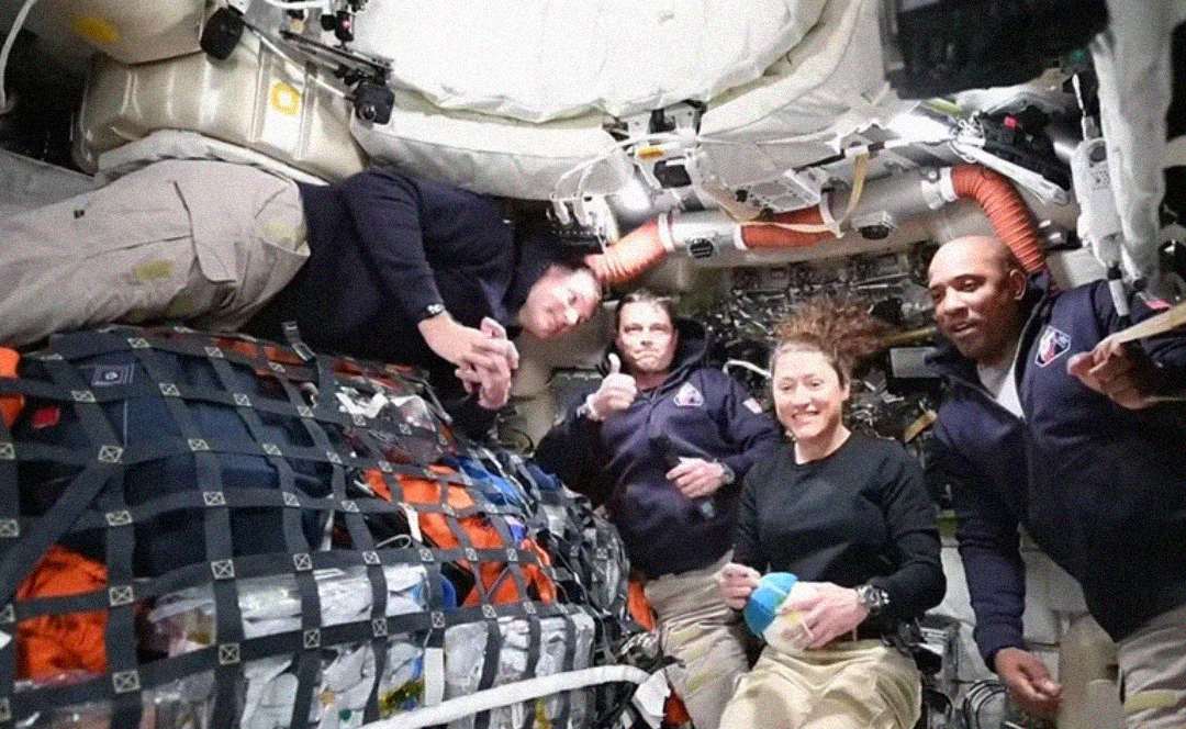 Four astronauts in casual uniforms float inside a crowded, equipment-filled spacecraft, smiling and posing for the camera in a zero-gravity environment.
