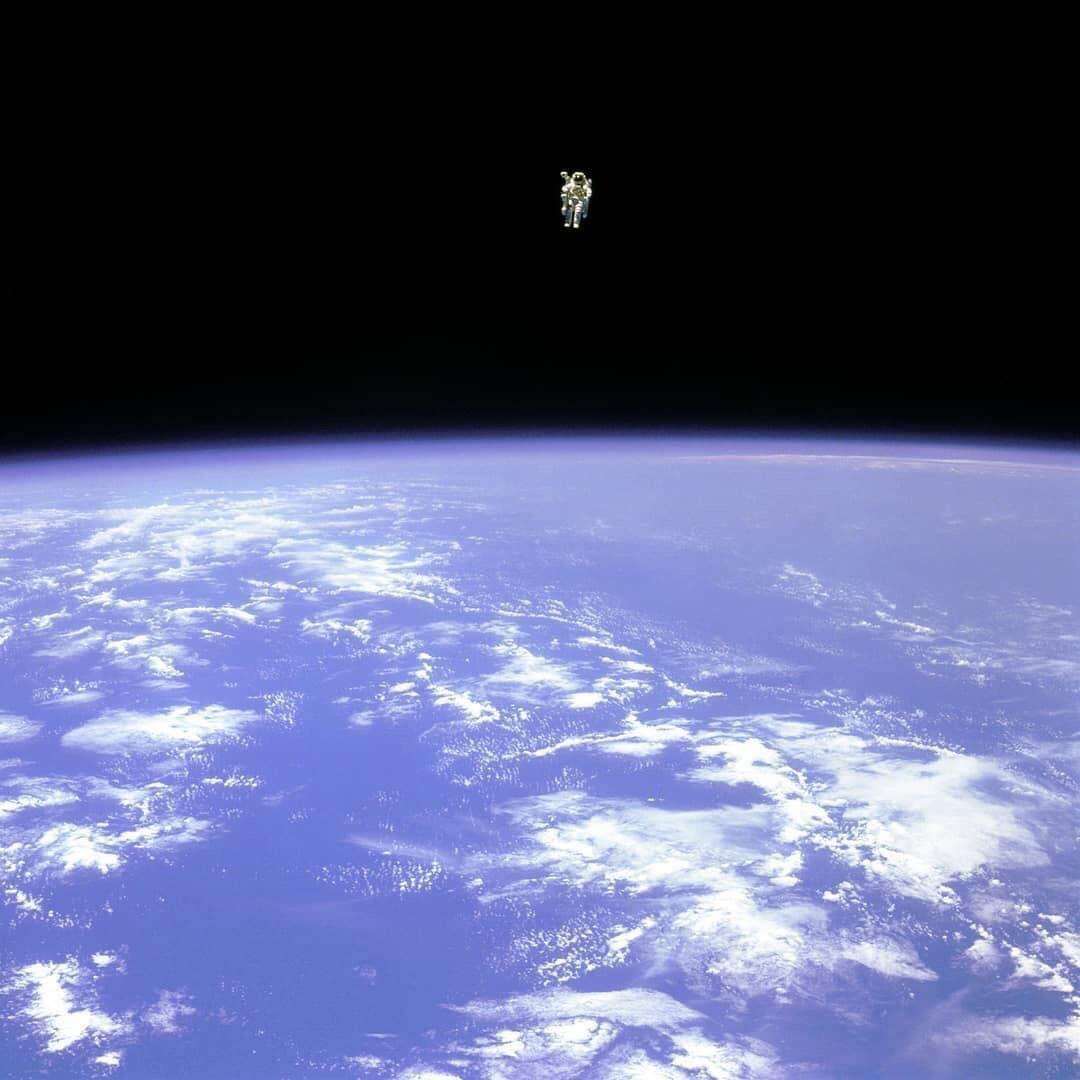 An astronaut floats alone in space above Earth, with the planet's blue surface and white clouds visible below and the vast blackness of space above.