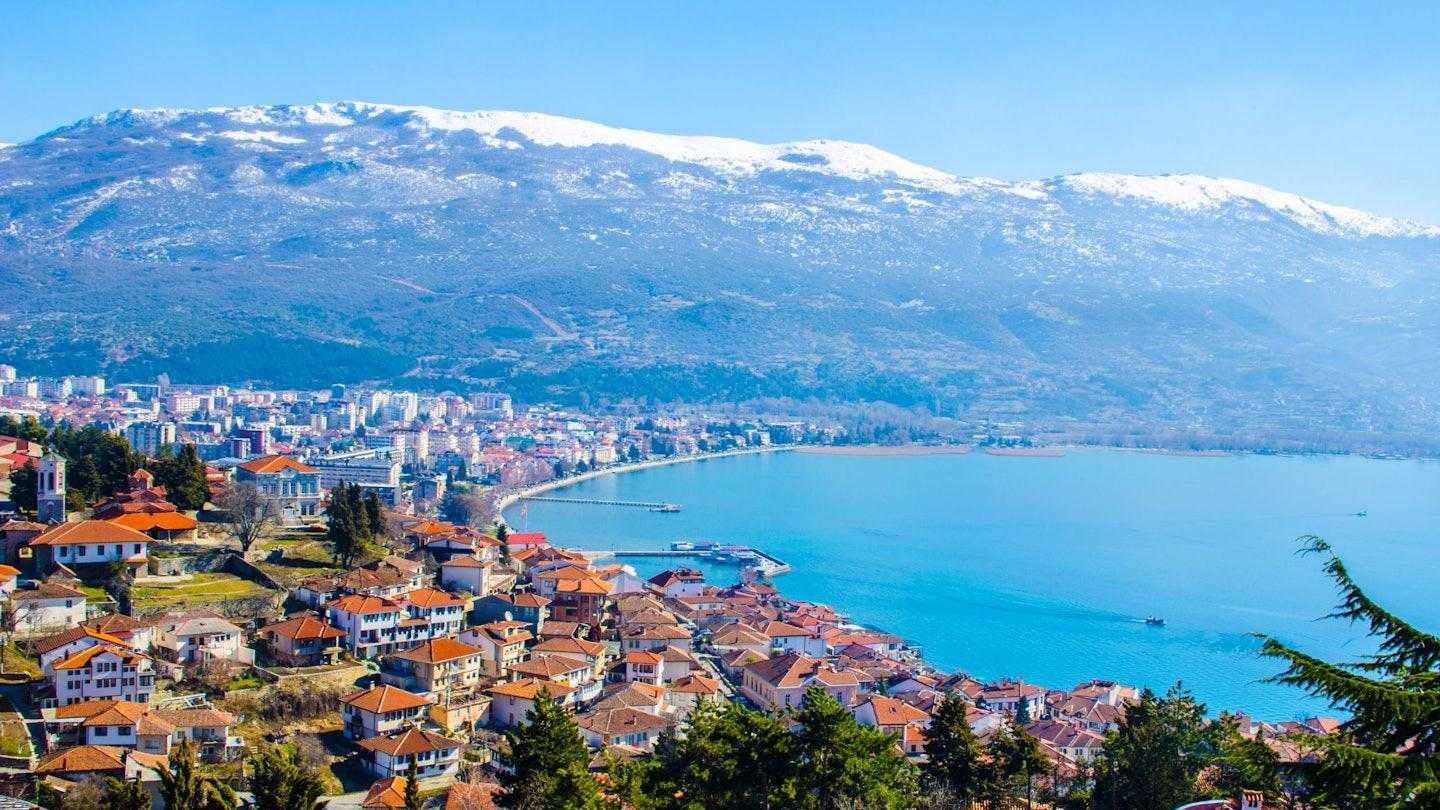 A scenic view of a lakeside town with red-roofed buildings, a calm blue lake, and snow-capped mountains in the background under a clear blue sky.