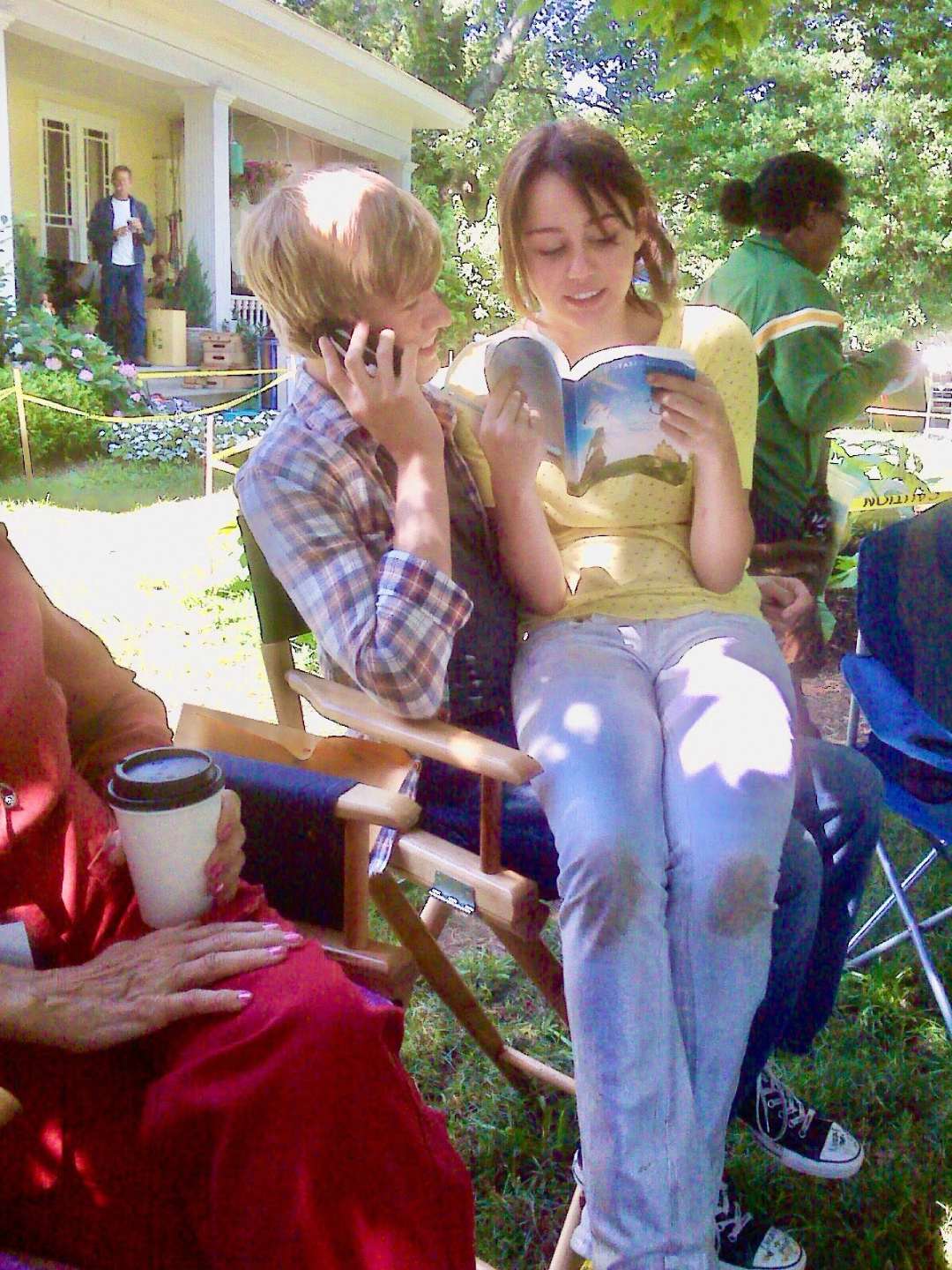 Two teenagers sit closely on a wooden chair outdoors; one is on the phone while the other reads a book. A person in red holds a coffee cup nearby, with a yellow house and other people visible in the background.