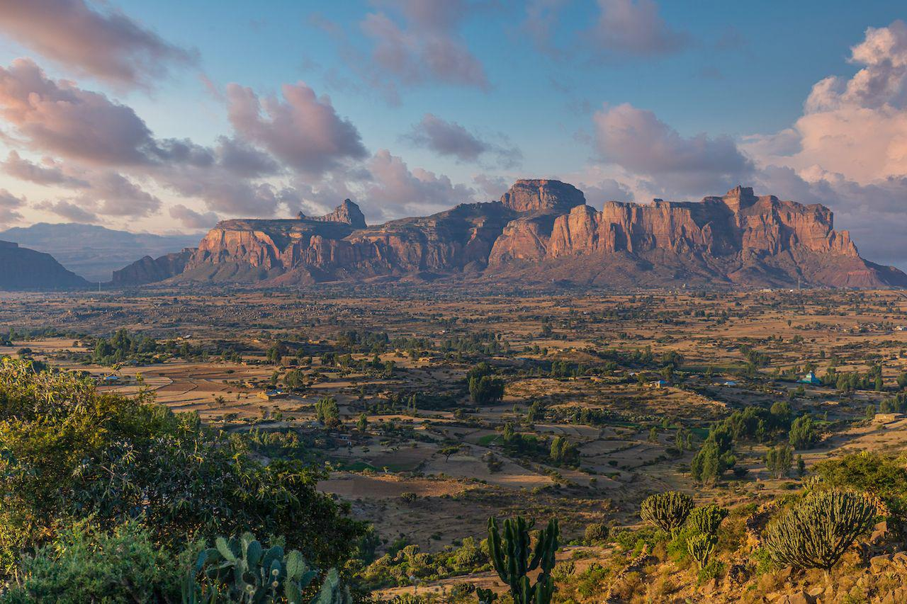 A wide view of the Gheralta Mountains in Ethiopia at sunset, with rugged red sandstone cliffs rising above green farmland and scattered trees under a partly cloudy sky.