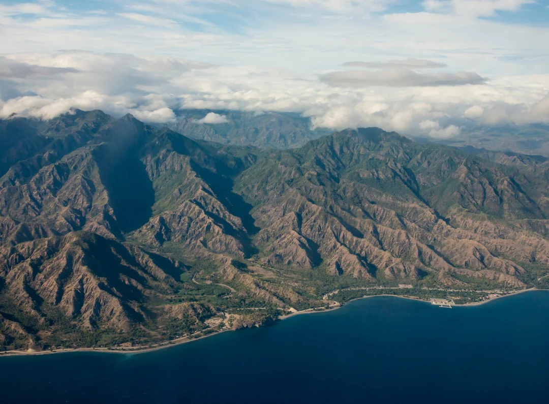 Aerial view of a rugged mountain range with green valleys, partially covered by clouds, meeting a calm, deep blue sea under a partly cloudy sky.