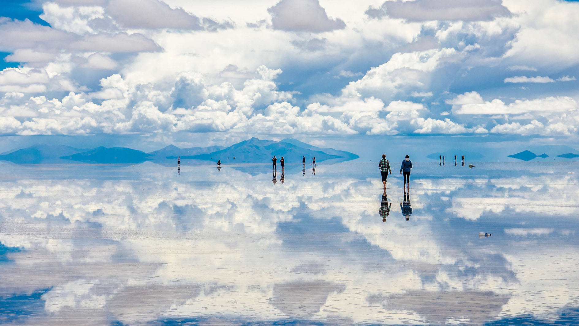 People walk across the reflective surface of Salar de Uyuni under a cloudy sky, creating a mirror effect that blends the sky and the horizon, with distant mountains visible in the background.