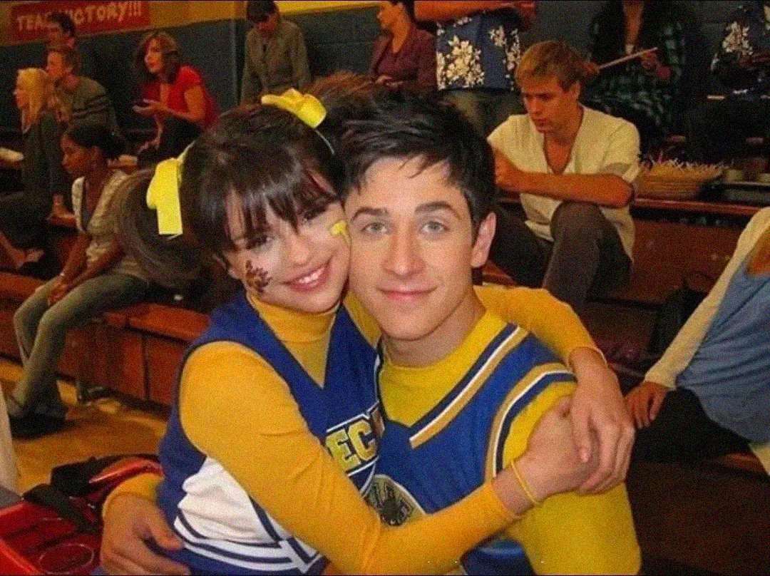 A girl in a cheerleader outfit with yellow pom-poms and face paint hugs a boy in a matching blue and yellow sports uniform. They are smiling and sitting in bleachers with other people in the background.