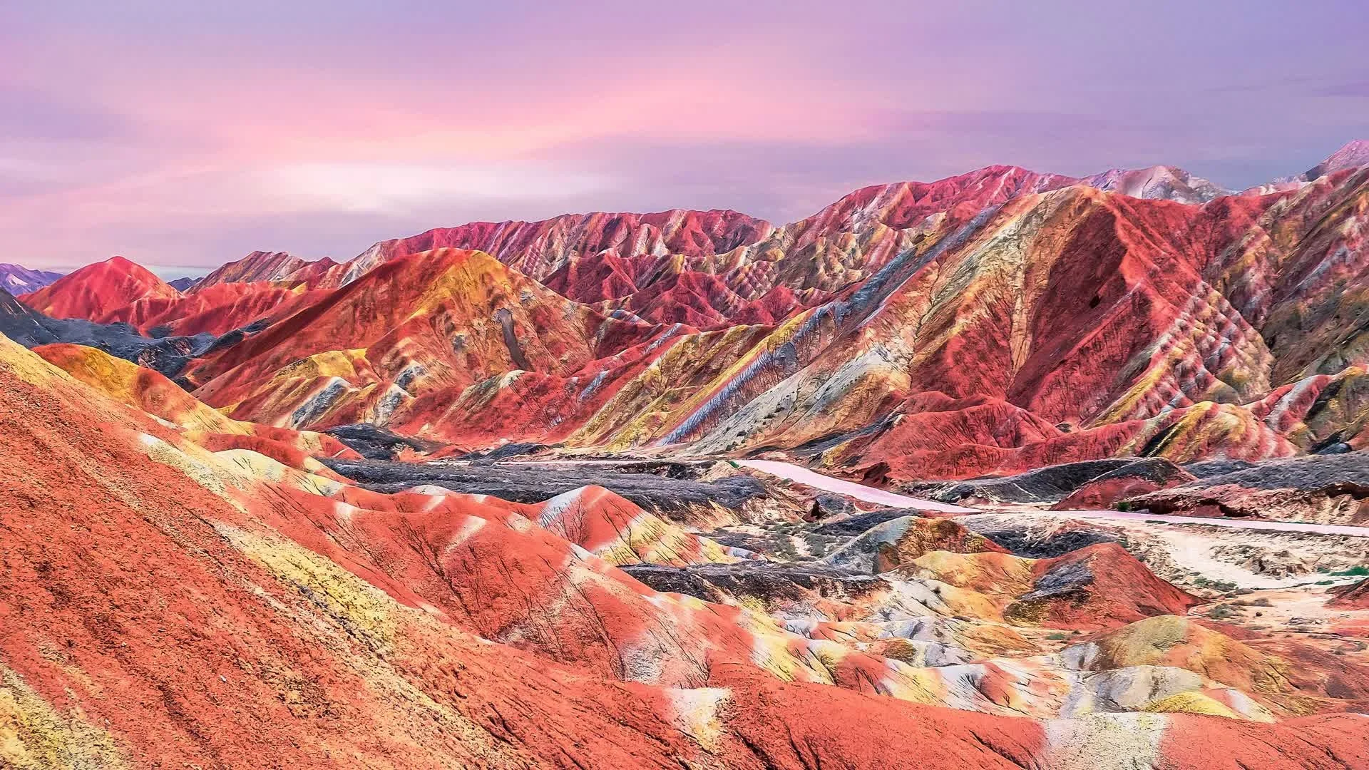 Colorful, layered hills and mountains with vibrant streaks of red, yellow, orange, and brown under a pastel pink and purple sky, showcasing a surreal landscape.
