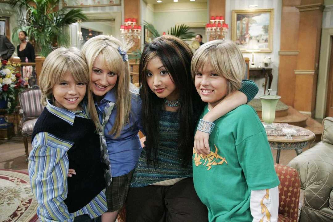 Four smiling teens stand together in a lavish living room, with two boys on either side of two girls. They pose closely, arms around each other, in a cheerful and friendly atmosphere.