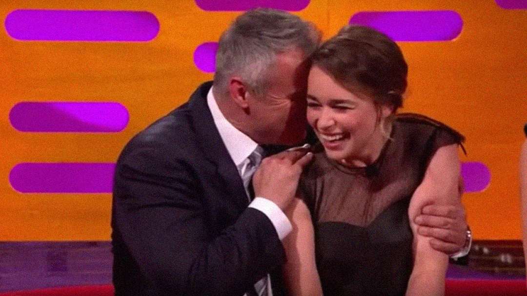 A man in a suit smiles and leans in to whisper to a woman in a black dress, who is laughing joyfully. They are sitting together on a talk show set with a bright orange and pink background.