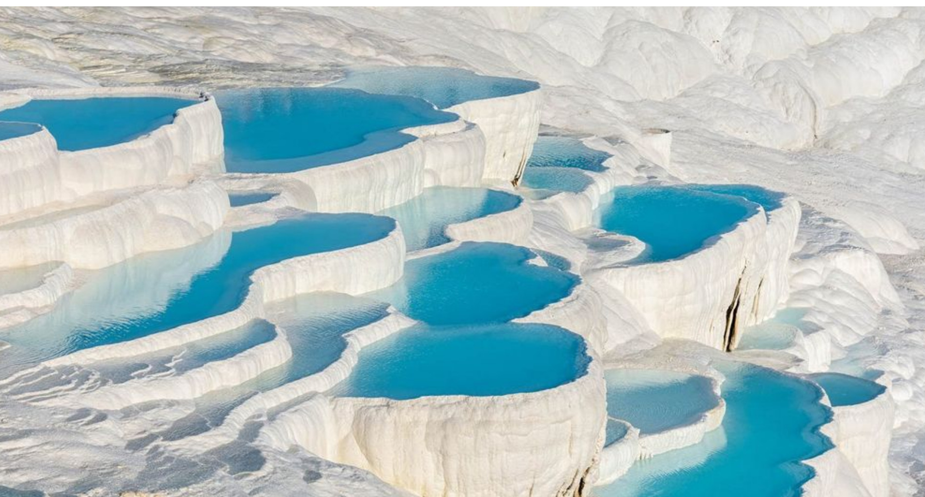 Terraced pools filled with bright blue water, surrounded by white mineral-rich formations, resembling steps. The scene looks natural and serene, with various pool levels and smooth, undulating white surfaces.