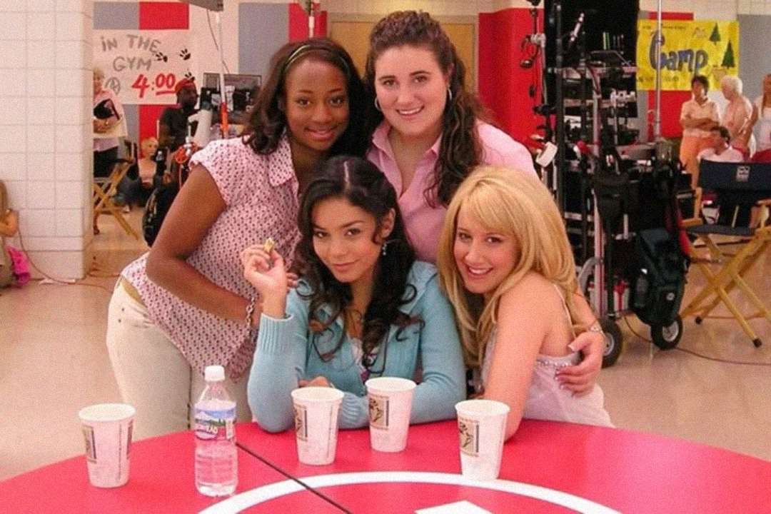 Four young women smile together in a school cafeteria setting, with three sitting at a red table with drinks and one standing behind them. Filming equipment and crew are visible in the background.