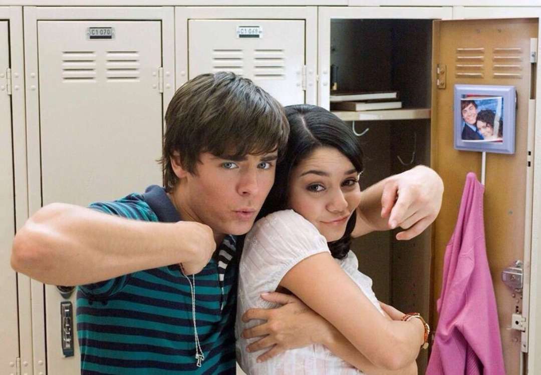 Two teenagers pose playfully in front of open school lockers. The boy points at himself while hugging the girl, who smiles with her arms folded. A framed photo and a pink jacket are visible inside the open locker.