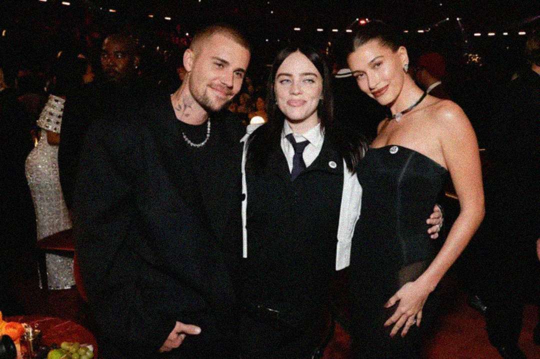 Three people pose together at a formal event. The person on the left wears a black suit and necklace, the person in the middle wears a black jacket over a white shirt and tie, and the person on the right wears a strapless black dress.