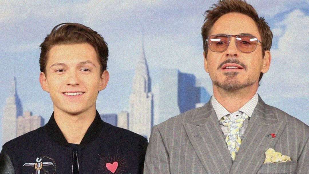 Two men stand side by side smiling; one wears a black jacket with heart and spider designs, the other wears a gray pinstripe suit, tie, and tinted glasses. A city skyline is blurred in the background.