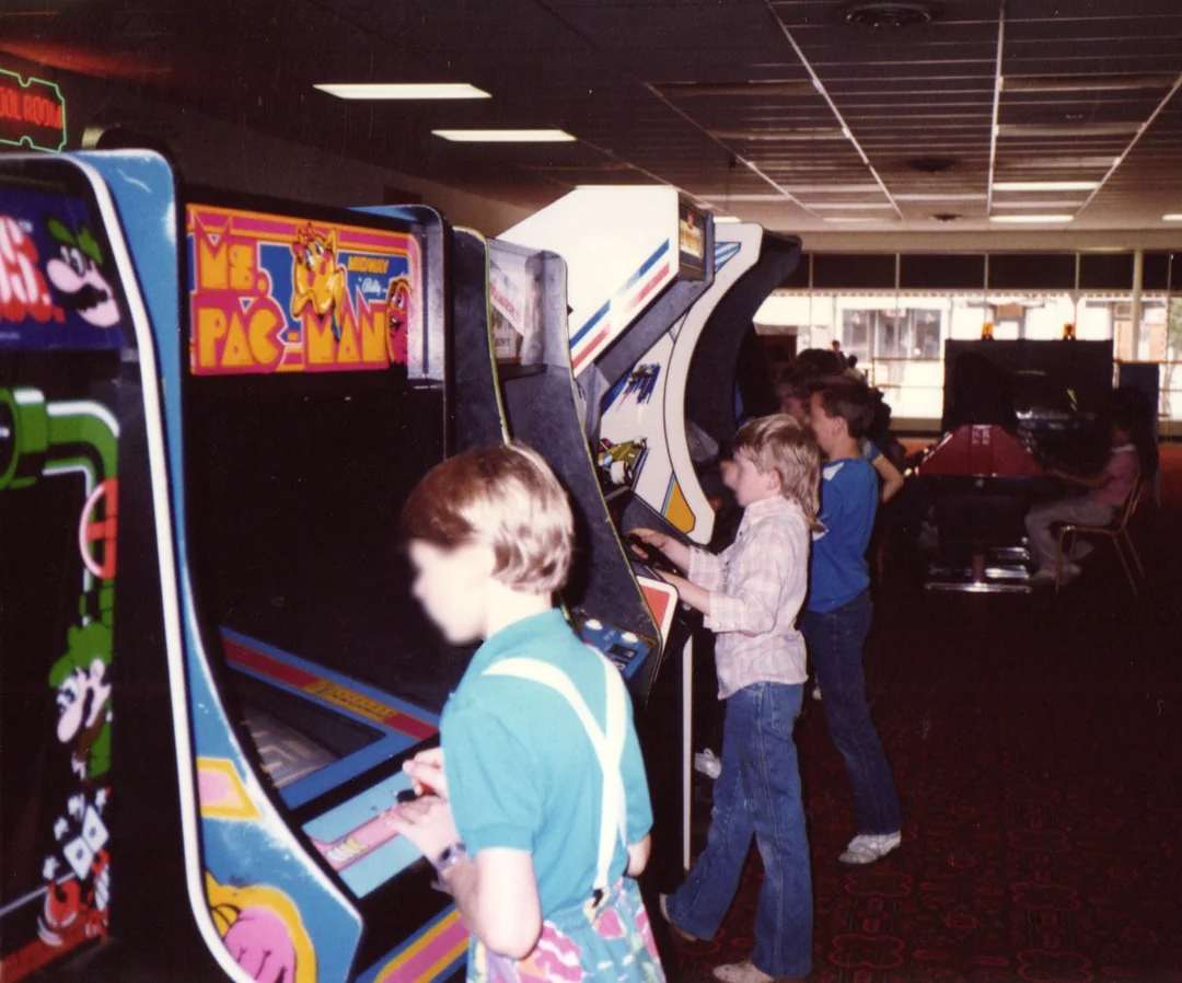 Children stand in a row playing vintage arcade games, including Ms. Pac-Man, in a dimly lit arcade with patterned carpet and large windows in the background.