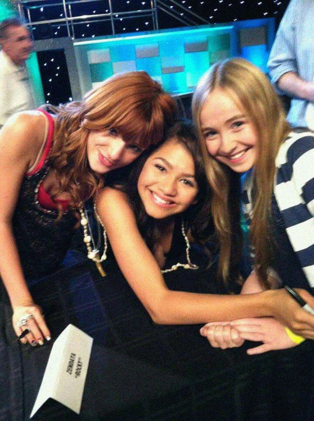 Three young girls smile for a photo together at a signing event. One is seated, while the other two lean in close. There are tables, a name placard, and a vibrant stage setup in the background.
