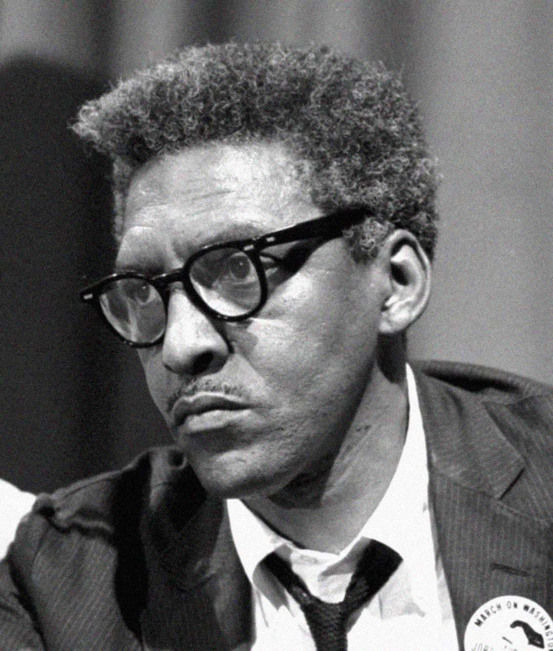 Black-and-white photo of a man with short curly hair, glasses, and a mustache, wearing a suit and tie with a button on his lapel, looking serious and thoughtful.