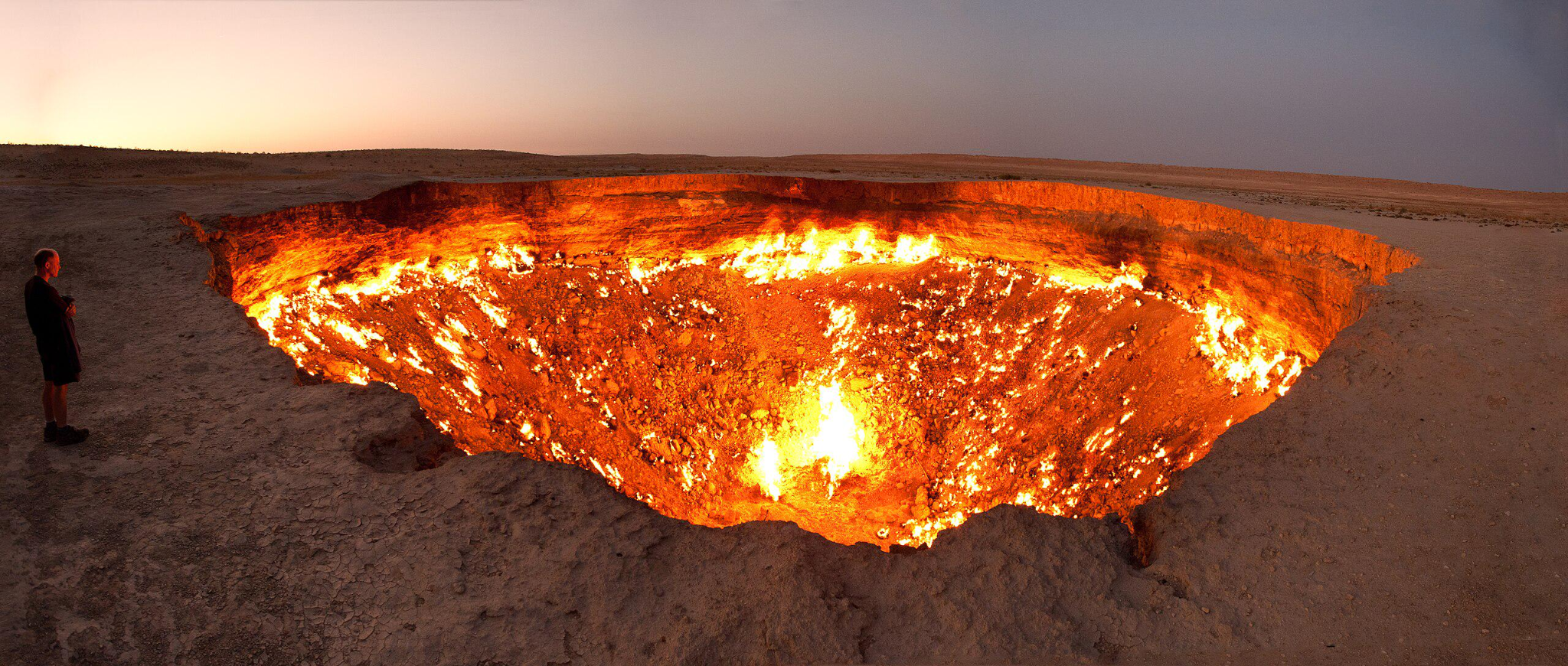 A person stands near the edge of a large, fiery crater in a barren, desert landscape at dusk, with intense flames and glowing embers illuminating the pit’s interior.